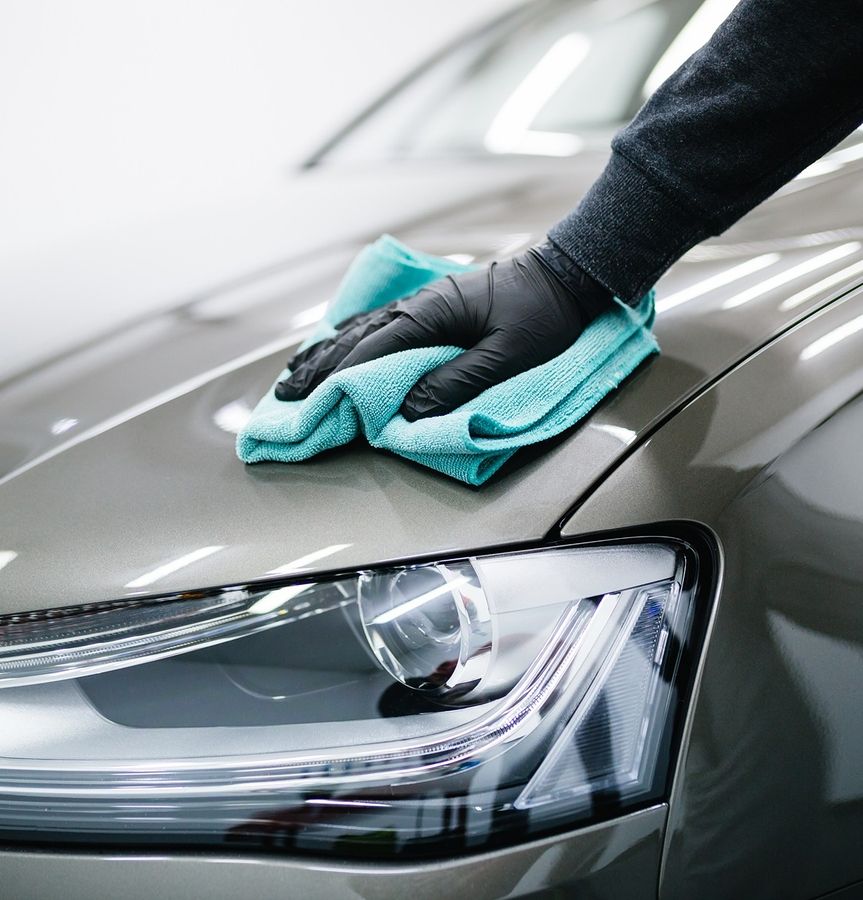 Hand mit schwarzem Handschuh wischt mit einem blauen Mikrofasertuch die Motorhaube eines silbernen Autos neben dem Scheinwerfer